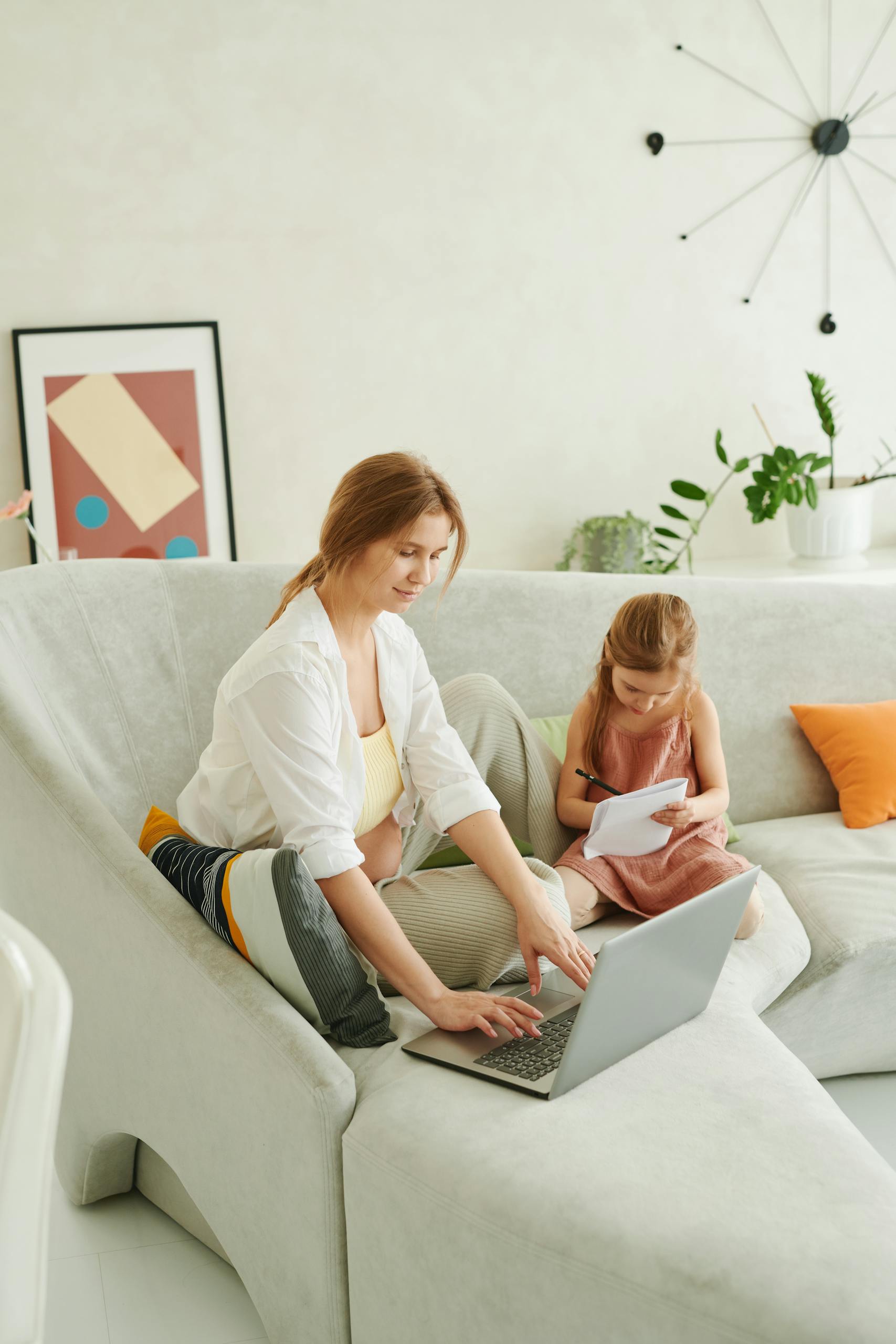 A mother working on a laptop while her daughter engages with a tablet in a cozy living room.