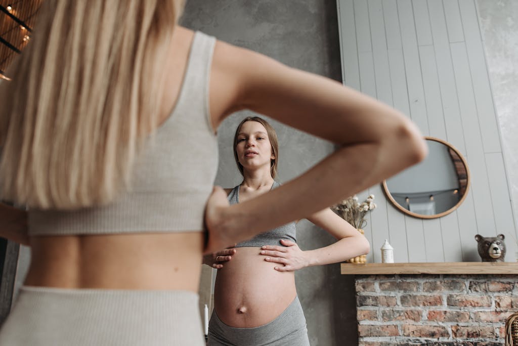 A pregnant woman practicing gentle exercise indoors, reflected in a mirror, in casual sportswear.
