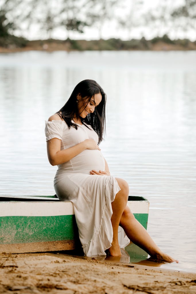 Expectant mother in a white dress sits by the beach, embracing maternity.